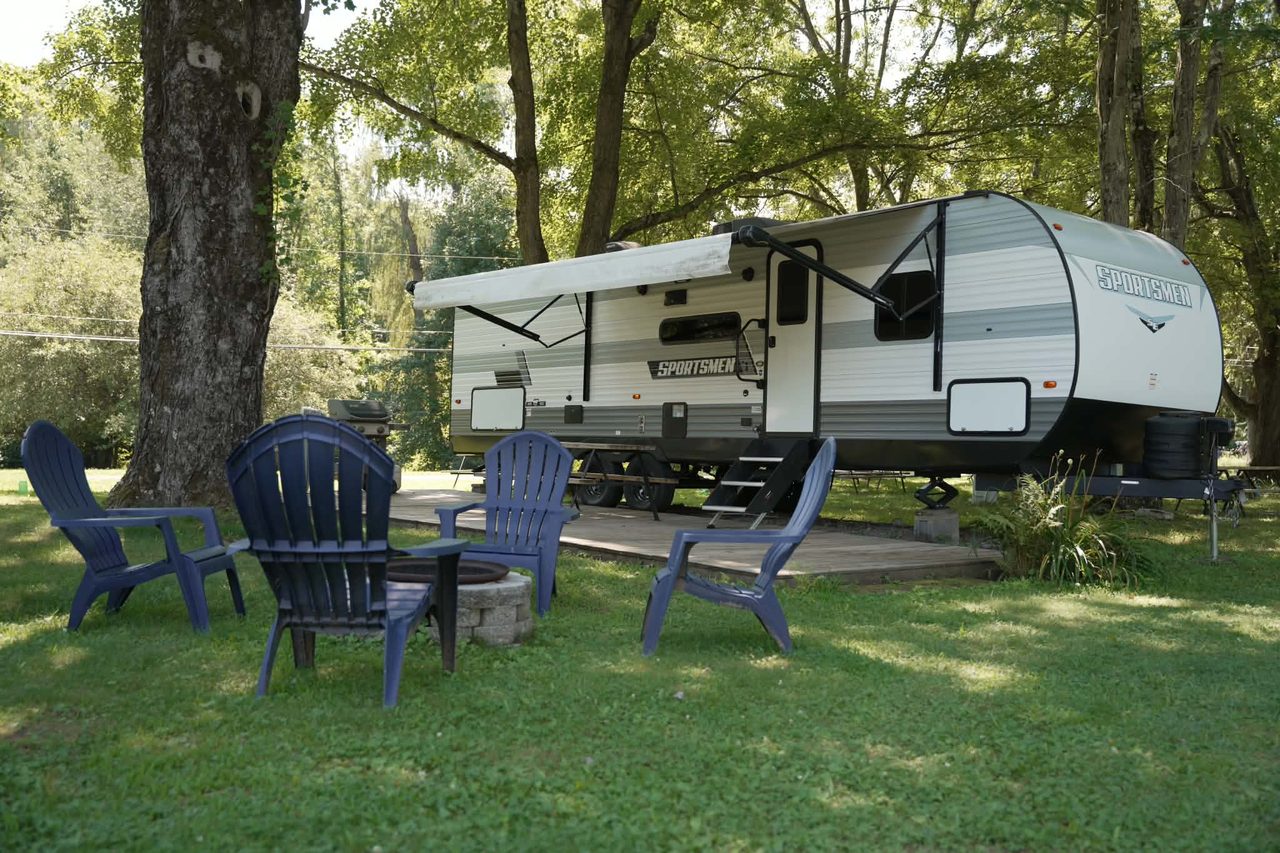 Bunkhouse RV rental with its awning out, Adirondack chairs and a fire pit under shade trees
